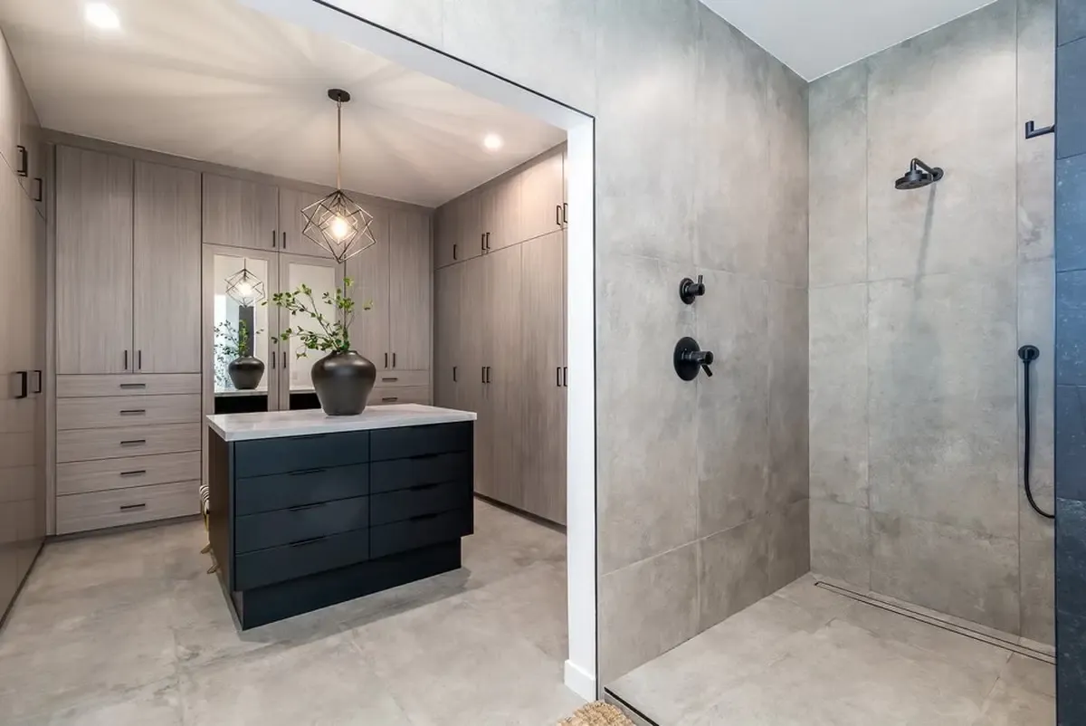 Modern minimalist bathroom and walk-in closet with concrete-look tiles, light wood cabinetry with black hardware, central island with black base, and open shower with black fixtures.