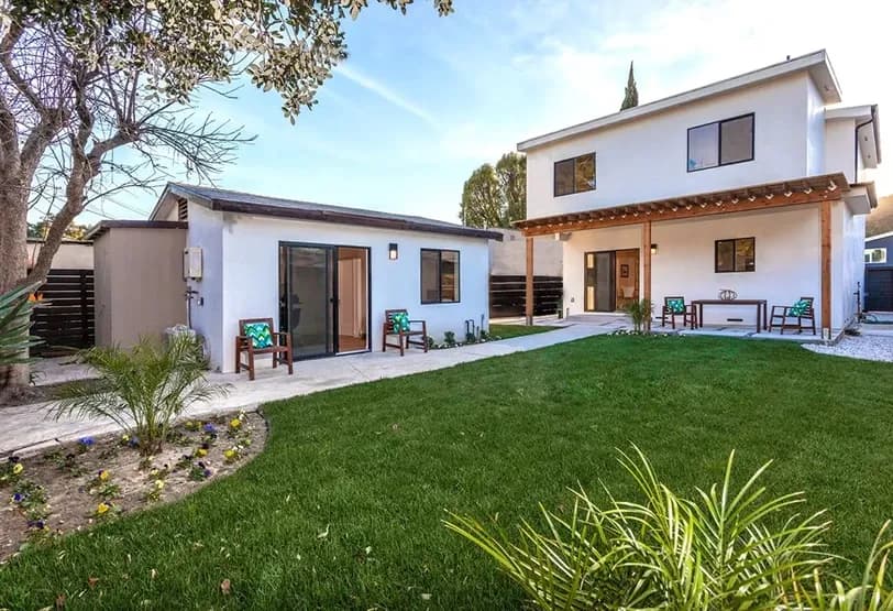 Modern two-story white residential property with well-manicured lawn, covered patio with outdoor seating, and adjacent single-story ADU with sliding glass doors.