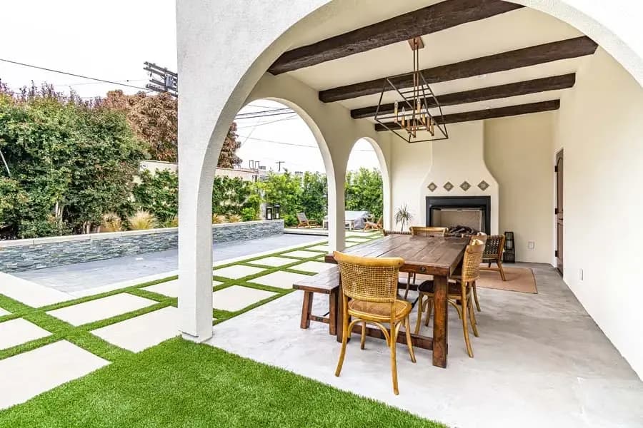Stylish outdoor living space with covered patio featuring white stucco walls and arched openings, wooden dining table with wicker chairs, and interior fireplace with decorative tile accents.