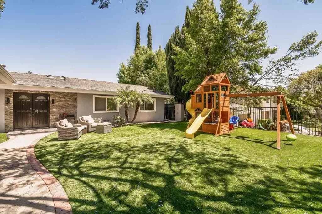 A backyard with a single-story house, green lawn, wooden playset with slide and swings, and patio furniture