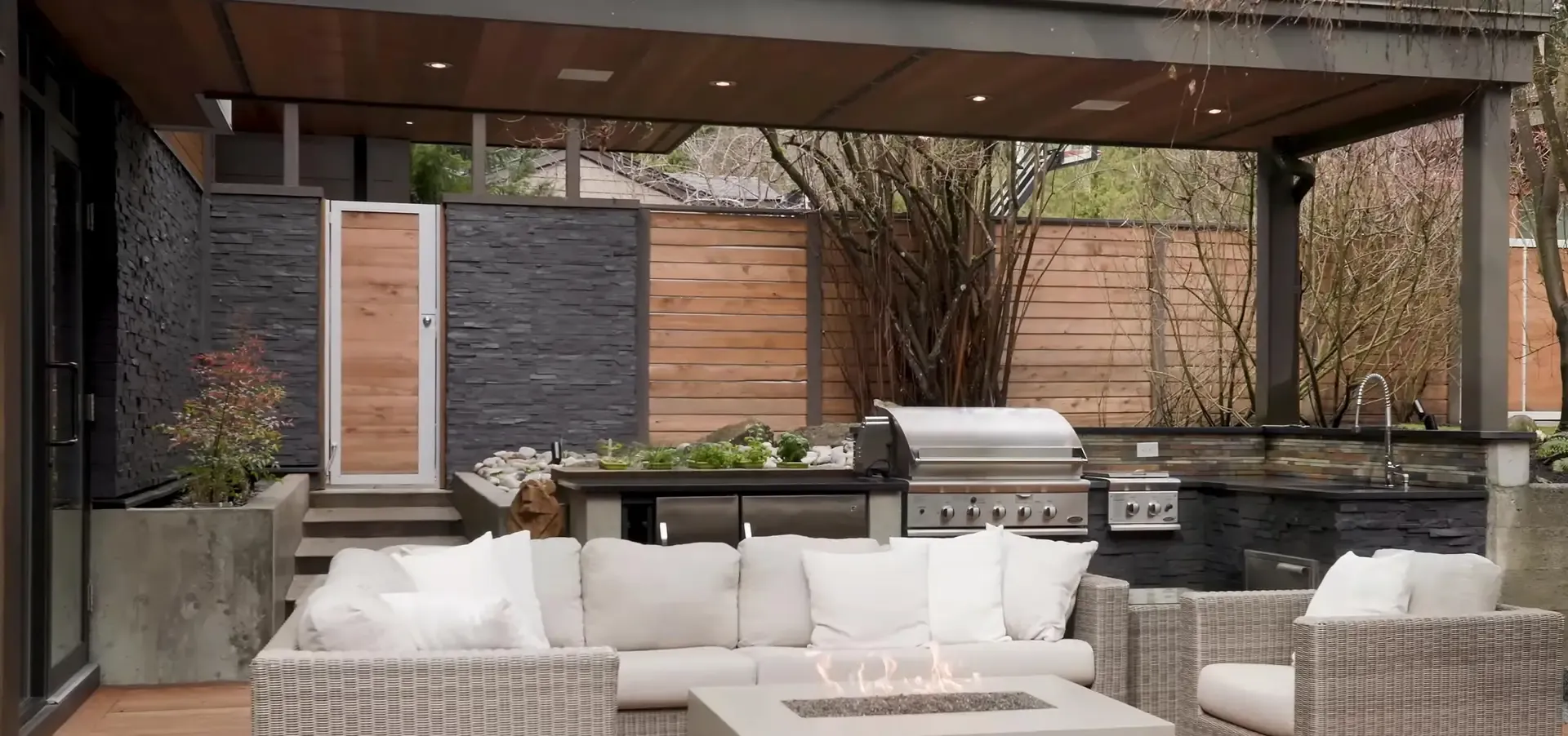 A covered outdoor patio with wicker furniture, white cushions, fire pit coffee table, and built-in outdoor kitchen