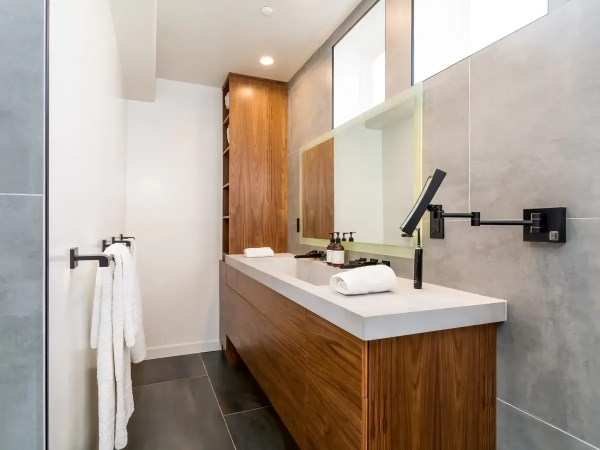 A modern, minimalist bathroom featuring a long vanity with light gray countertops and warm wood cabinetry, large mirrors, and light gray wall tiles.