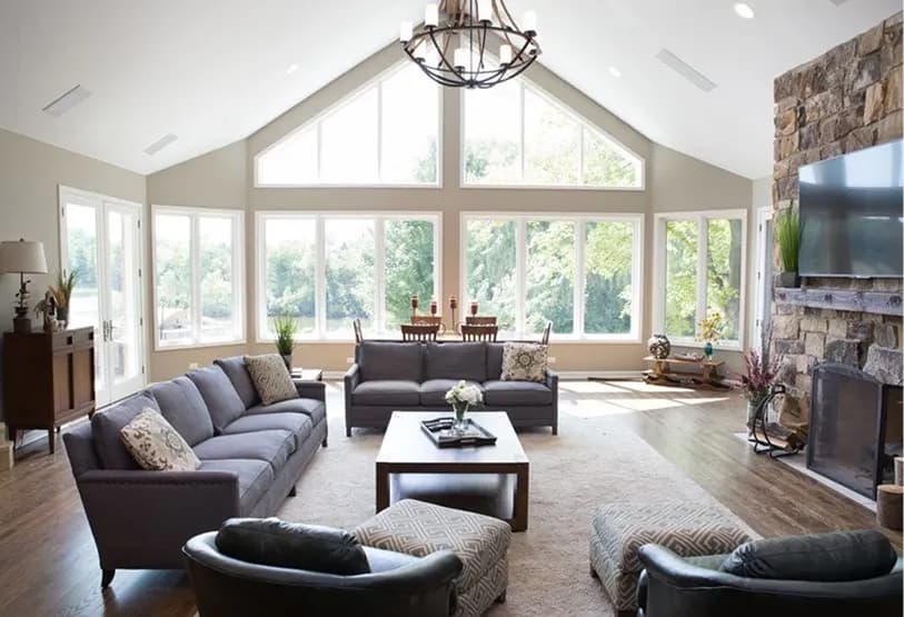 A spacious, bright living room with high ceilings and large windows offering views of greenery, featuring two gray sofas, dark leather chairs, and a stone fireplace.
