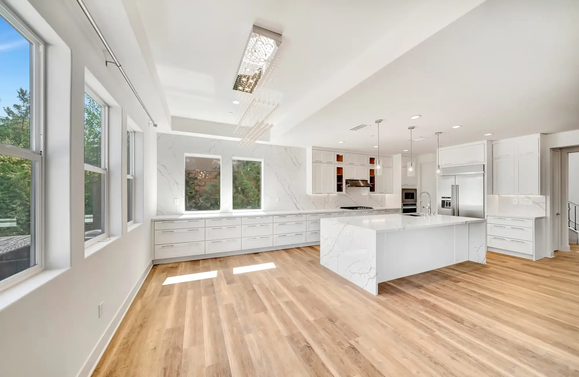 A spacious, modern kitchen with a large central island featuring a white marble countertop, white cabinetry, stainless steel appliances, and light wood flooring.