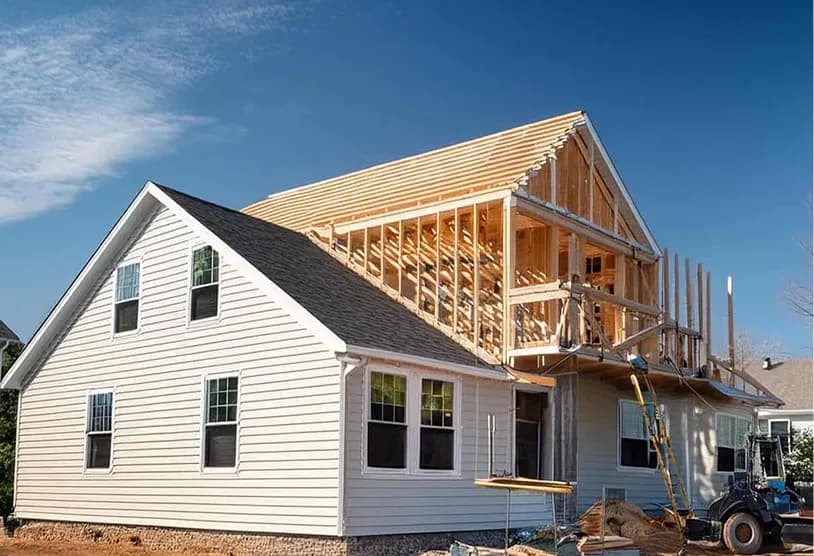 A residential house undergoing renovation with a second story addition being constructed. The existing house has white siding and a dark shingled roof with the new upper level visible in its wooden framing stage.