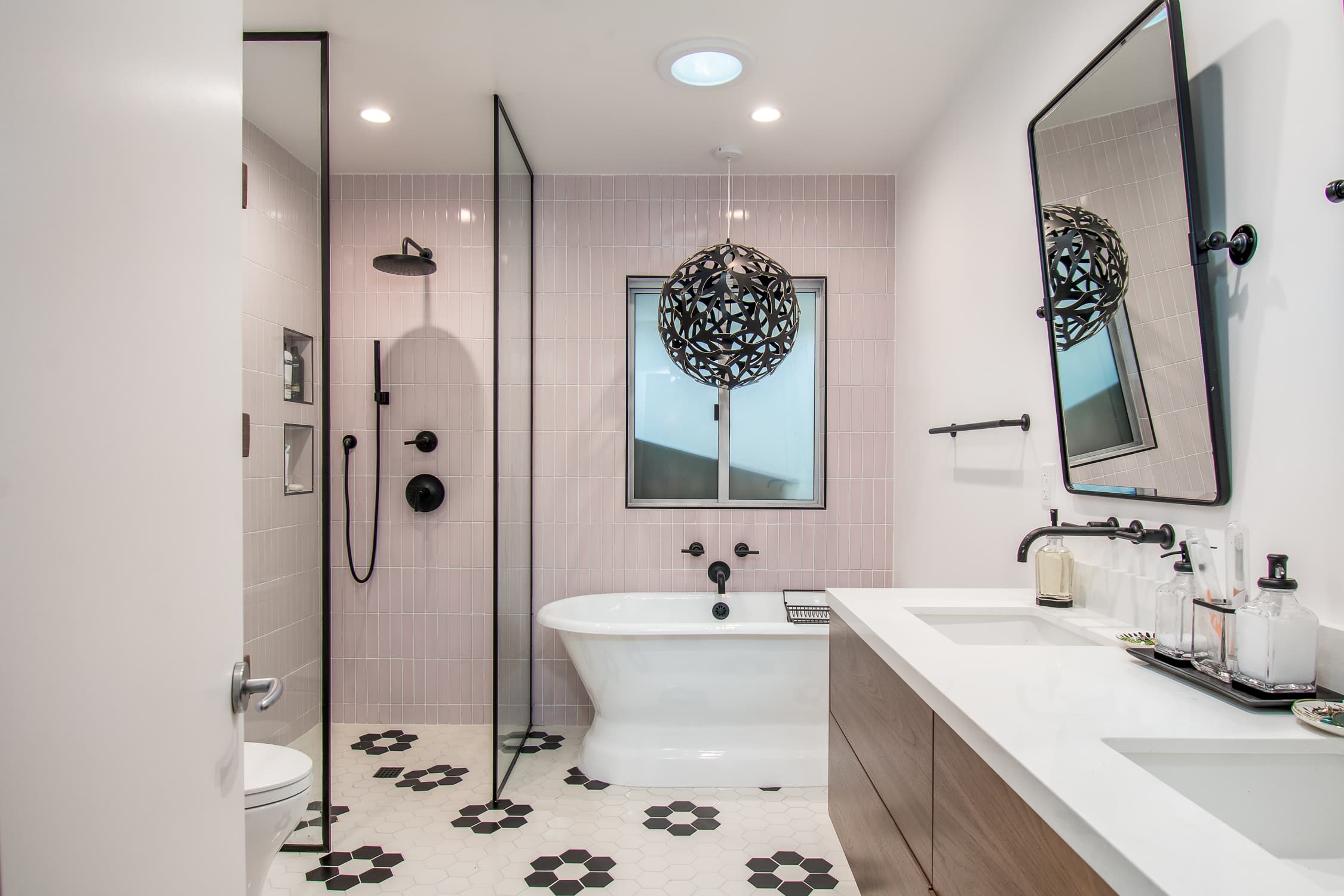 An elegant bathroom with soft pink wall tiles and white hexagonal floor tiles with black accents. Includes a white freestanding clawfoot bathtub, walk-in shower with black fixtures, double vanity with wood cabinets, and a unique black spherical pendant light.