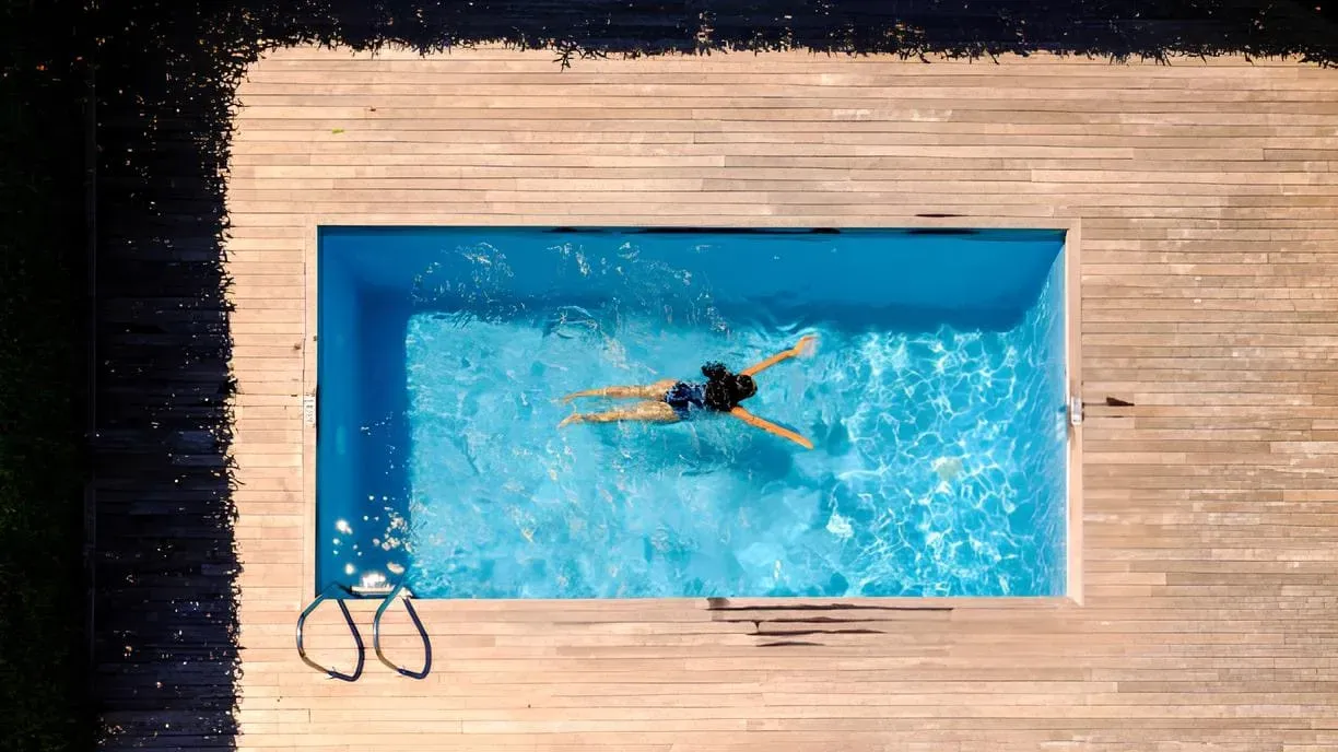 An aerial view of a rectangular swimming pool with bright turquoise water surrounded by a light wooden deck. A single swimmer performs laps in the center of the pool, with trees bordering the area on a sunny day.