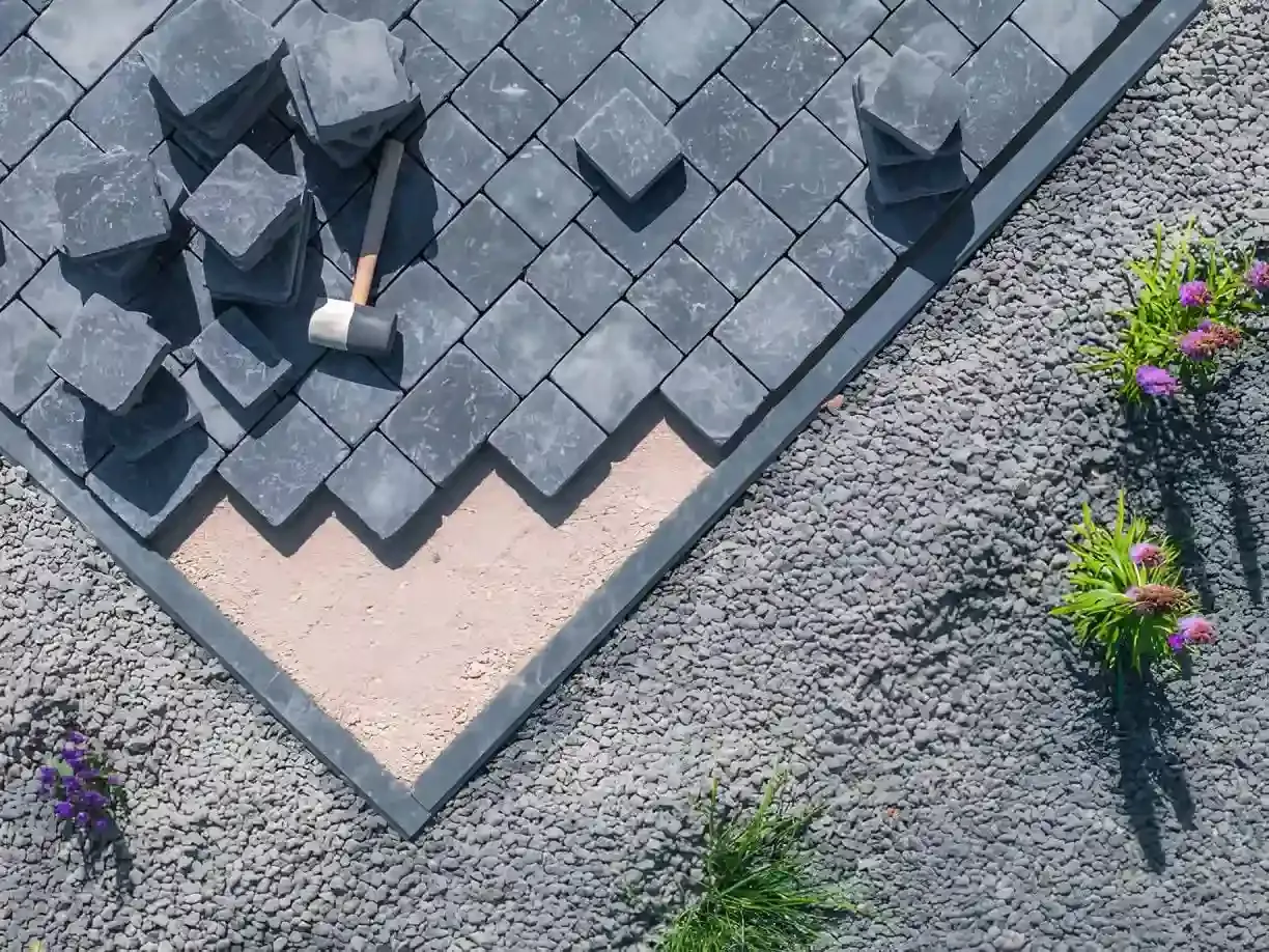 A top-down view of paver stone installation in progress, showing dark gray rectangular stones in a herringbone pattern, surrounded by gravel border, with sand base and a rubber mallet indicating active construction.