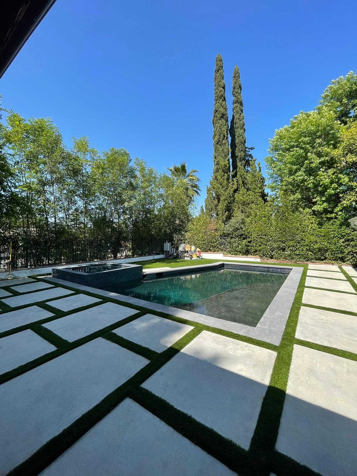 A newly remodeled backyard with a rectangular dark turquoise pool, adjacent square spa, geometric patterned patio with light gray pavers and dark green artificial grass strips, and tall Italian cypress trees in the background.