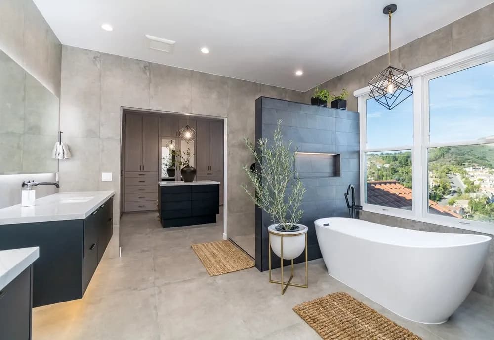 A luxurious bathroom with a freestanding oval tub, double vanity with dark gray cabinets, and a walk-in shower. Large windows overlook a residential neighborhood, with geometric pendant lighting and potted plants adding style.