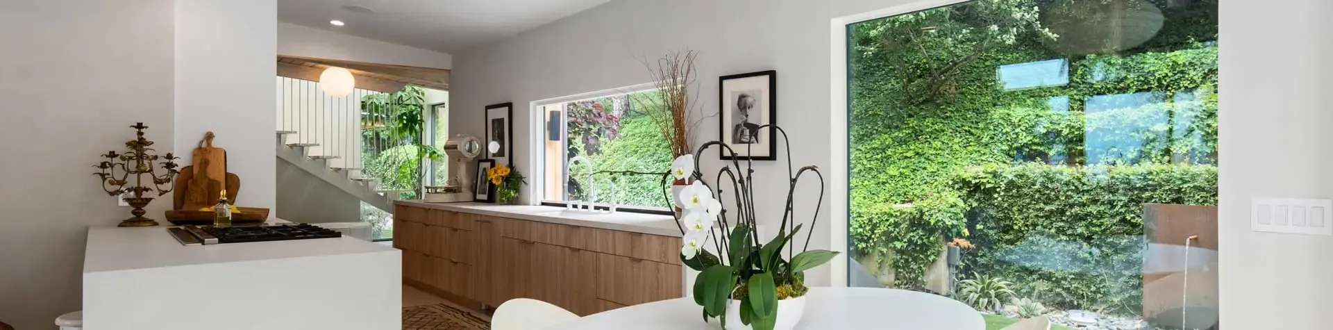 Bright airy modern kitchen and dining area with light wood cabinetry, white countertops, and large window opening to lush green outdoor garden.
