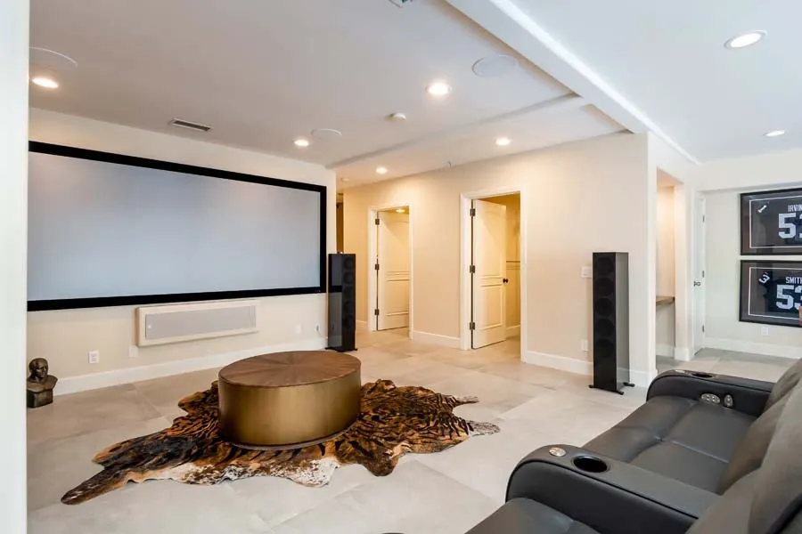 Modern home theater or media room with large wall-mounted projection screen, black leather reclining sofa, gold coffee table on animal hide rug, and tall black speakers.