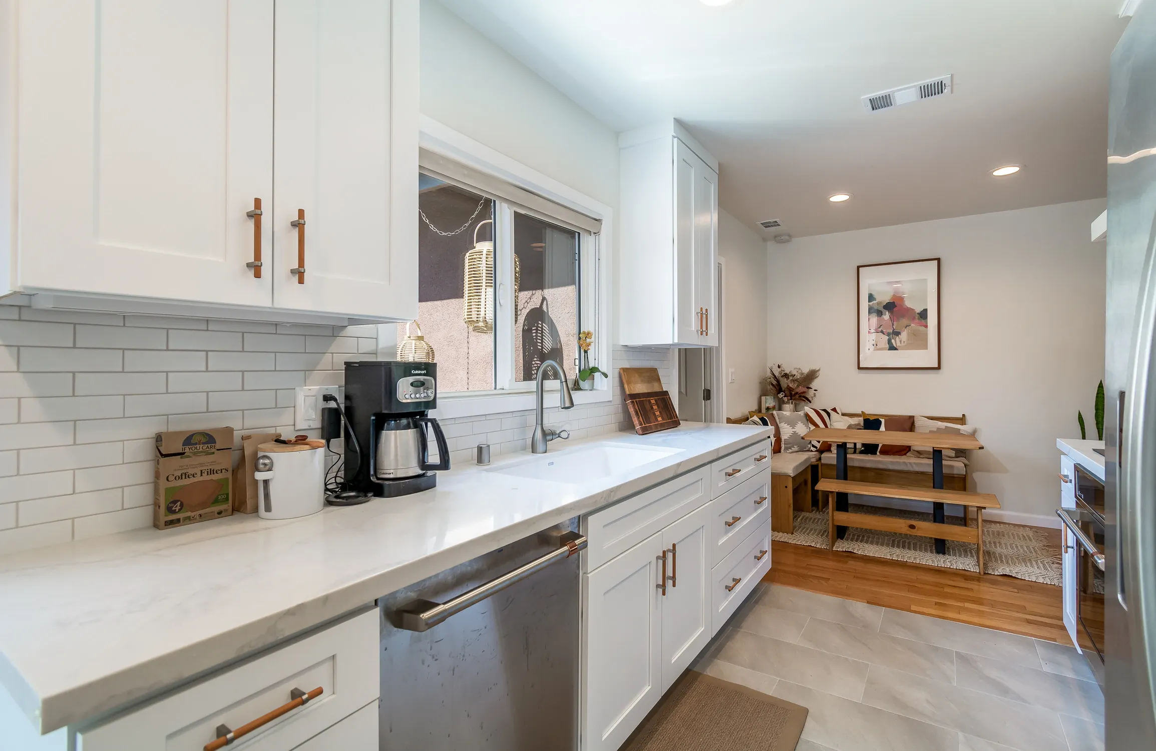 An open-concept kitchen with white cabinetry, subway tile backsplash, wooden dining table with benches, and a window above the sink