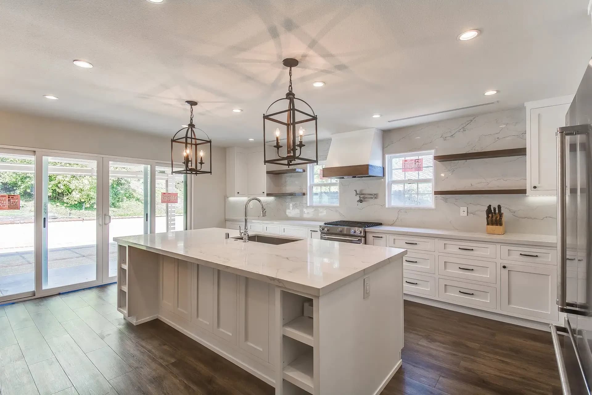 A bright, modern kitchen with white cabinetry and a large marble-topped island featuring a sink. The space includes stainless steel appliances, a gas range with a matching hood, and open shelving above the counter.