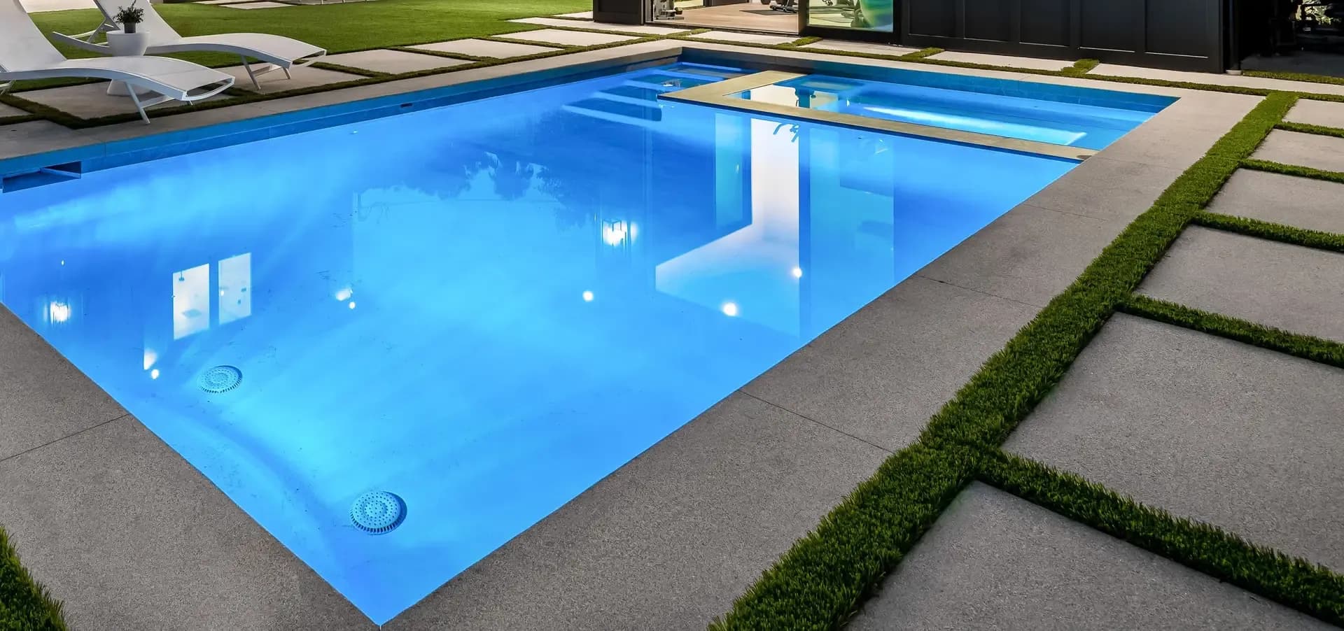 A modern rectangular swimming pool with clear blue water, surrounded by a light gray concrete deck with two white lounge chairs and a contemporary building in the background.