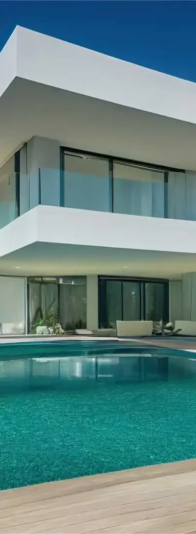 A modern, two-story house with a crisp white exterior and clean angular lines, featuring large floor-to-ceiling glass windows with dark frames. The house has a second-floor balcony with sleek glass railings. In the foreground, a rectangular swimming pool with vibrant turquoise water sits on a light wooden deck, reflecting the house and sky.