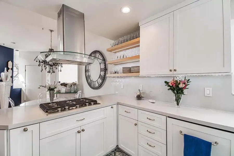 A modern, elegant white kitchen with stainless steel appliances, open shelving with glassware, a round black wall clock, patterned black and white floor tiles, and fresh pink flowers. The space features a gas cooktop with a sleek range hood and bright, airy atmosphere.
