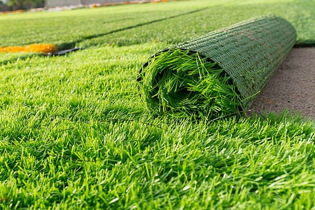 A large installation of bright green artificial turf grass with a rolled section in the foreground showing the synthetic blades and dark green woven backing. The scene depicts an outdoor installation site.