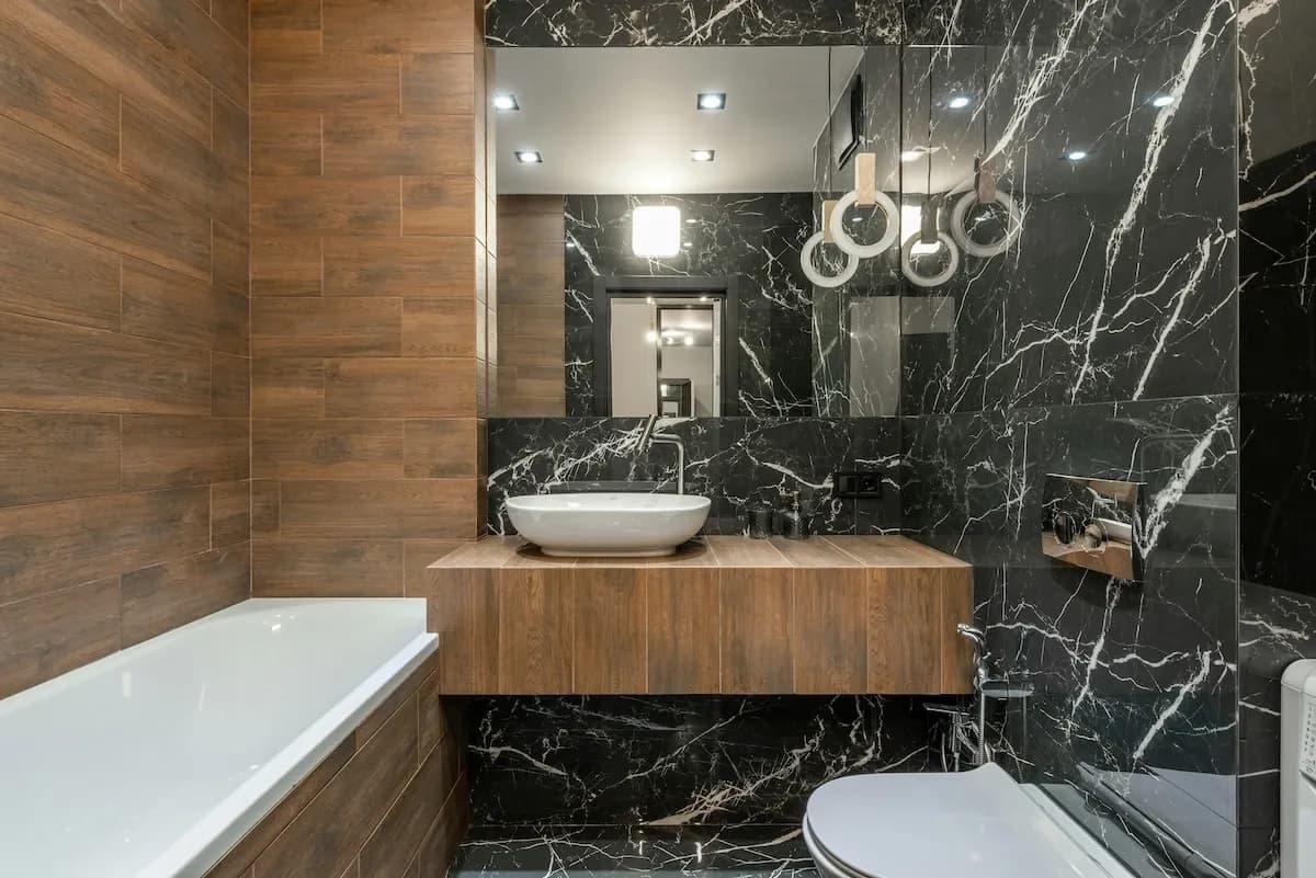 A modern bathroom with vertical warm wood paneling contrasted against glossy black marble tiles with white veining. Features a white floating bathtub, vessel sink, modern silver faucet, and unique ring-shaped pendant lights.
