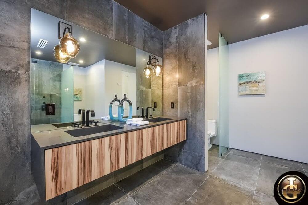 A contemporary industrial-style bathroom featuring a floating wood-grain double vanity with dark gray countertops, two vessel sinks with black faucets, frameless mirror, golden spherical wall sconces, and gray concrete-like tiled walls.
