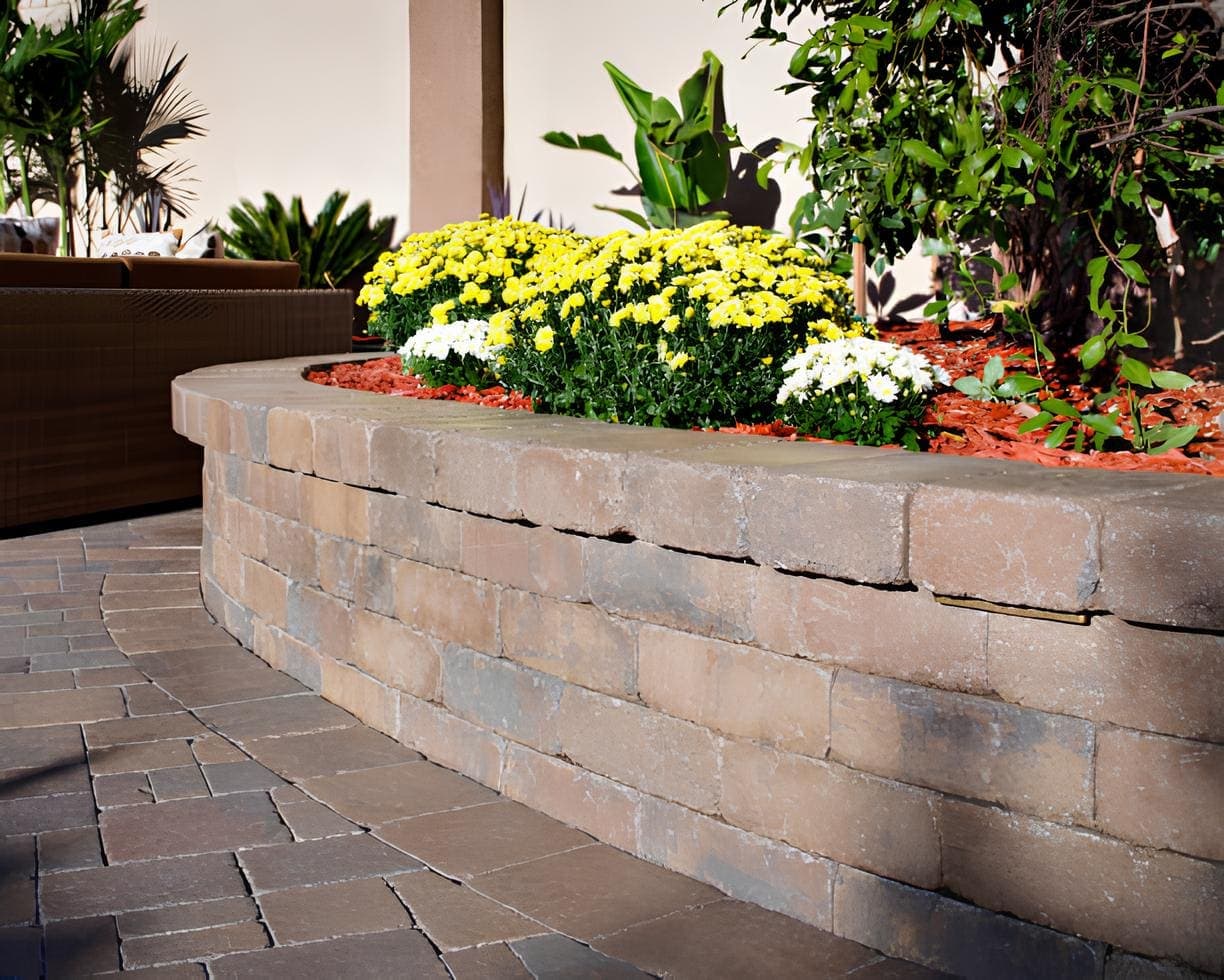 A beautifully maintained outdoor patio featuring a curved stone planter filled with vibrant yellow and white chrysanthemums, set against dark brown herringbone pavers, with wicker outdoor furniture and lush tropical plants.