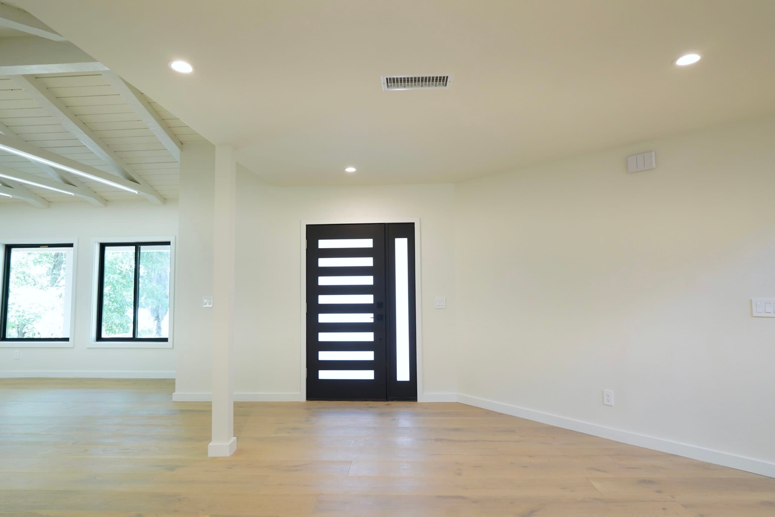A modern empty living room with light hardwood floors, cream-colored walls, and exposed white wooden ceiling beams. Features a striking black door with frosted glass panels, large black-framed windows overlooking greenery, and recessed lighting.