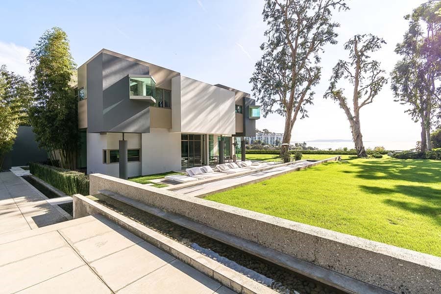 A luxurious modern multi-level house with gray and white exterior, floor-to-ceiling glass windows, and an expansive manicured lawn leading to ocean views. Features a sunken patio with lounge chairs and bamboo trees.