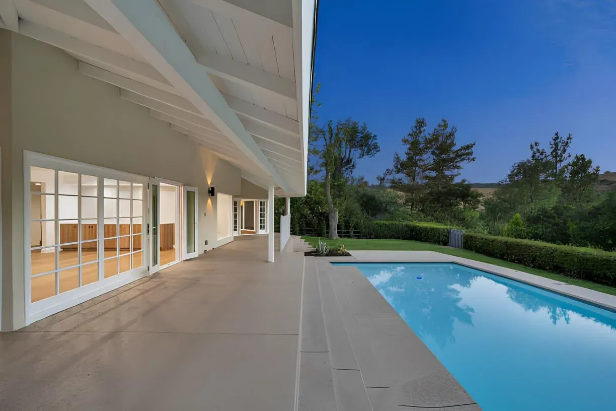 A luxurious outdoor living space with a modern house featuring beige exterior and white beams, adjacent to a turquoise infinity pool with light gray stone coping. Includes wooden lounge chairs with white cushions and a striped umbrella on a concrete patio.