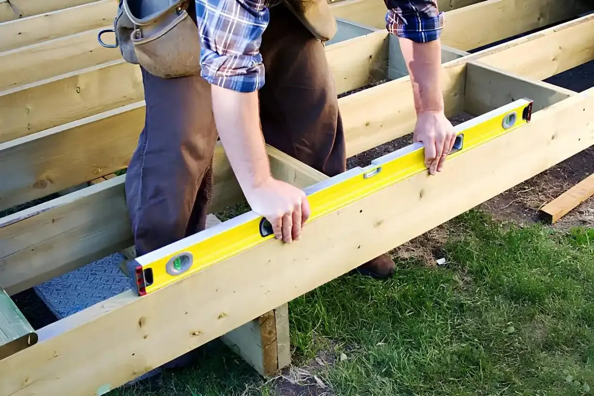 A construction worker wearing a blue and white plaid shirt using a yellow spirit level to check a horizontal wooden beam. The deck framing sits on blue concrete blocks with grass surroundings and natural wood materials.