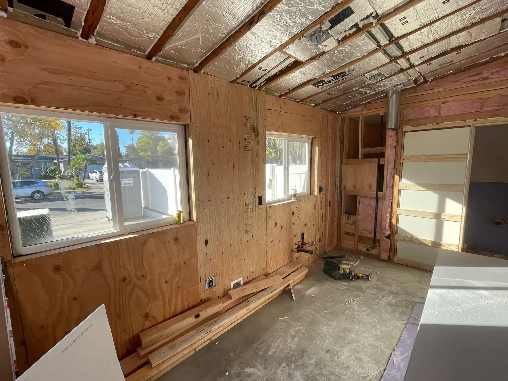 The interior of a room under construction with exposed plywood walls, bare concrete floor, and ceiling with insulation. Features two windows looking out to a street, with construction tools, lumber, and pink fiberglass insulation visible.