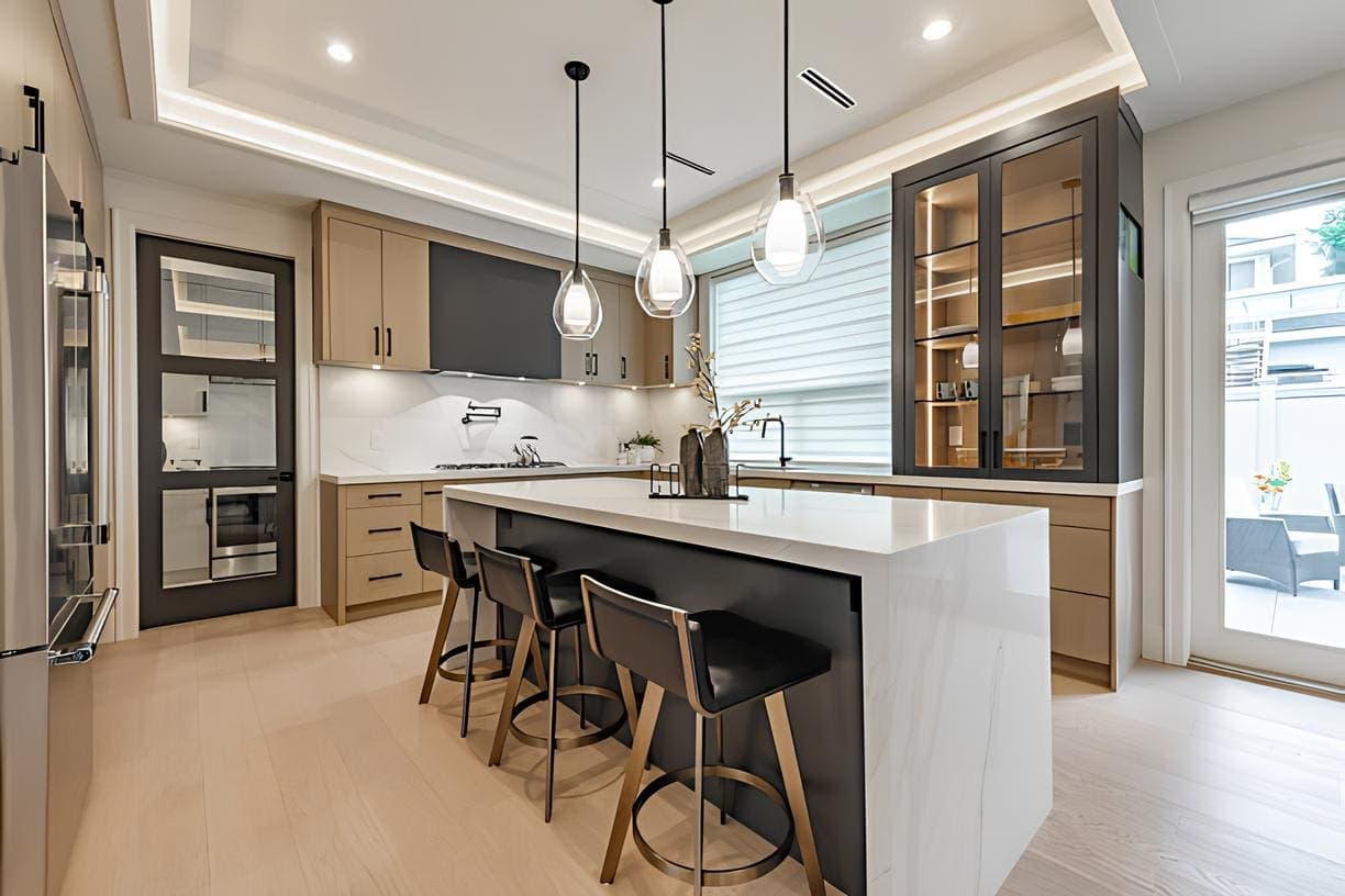 A modern luxury kitchen with a central white marble island, dark gray cabinetry, and light wood lower cabinets. Features stainless steel appliances, three pendant lights with glass shades, bar stools with gold bases, and a window with white blinds overlooking an outdoor patio.