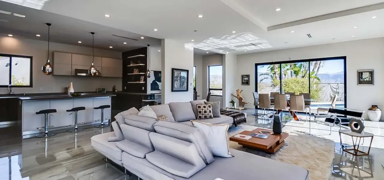 A modern, spacious open-concept living area with high ceilings, sliding glass doors opening to a pool with mountain views, a sectional sofa, and a kitchen with two-tone cabinetry.