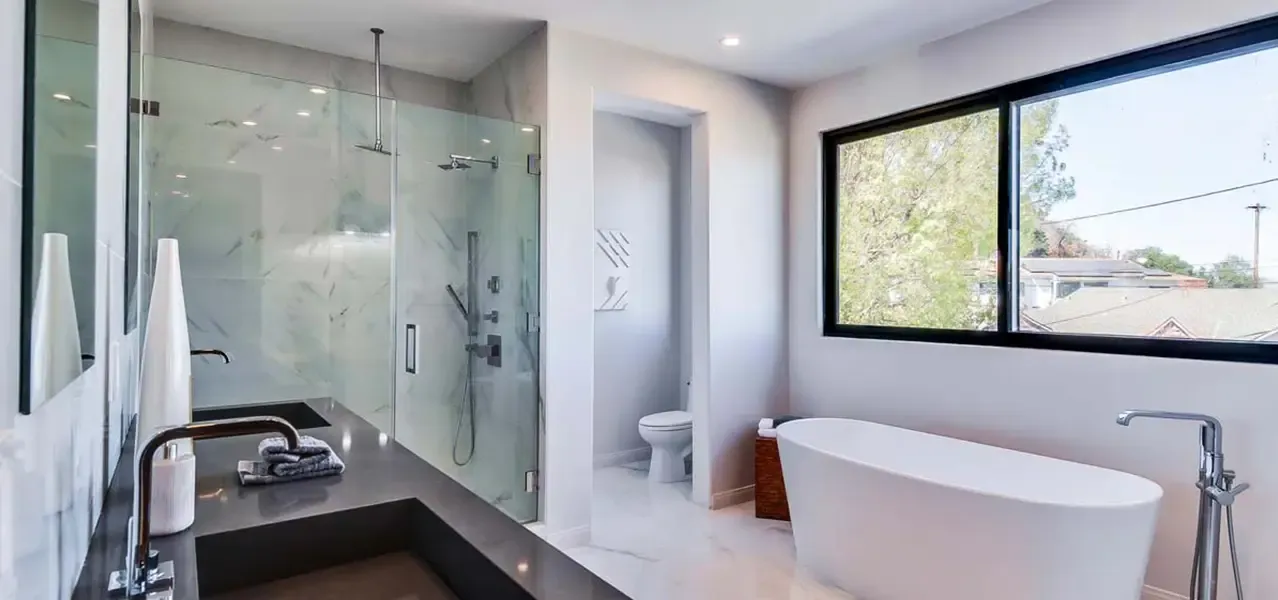 A modern luxury bathroom with a freestanding white oval bathtub, glass-enclosed shower with rain-style showerhead, dark countertop vanity with dual sinks, and large window with natural light.