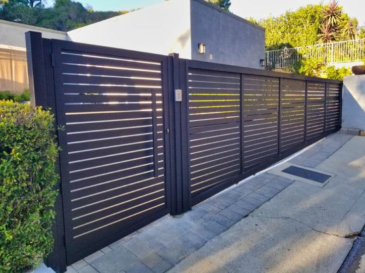 A modern black iron gate with horizontal slats at a property entrance, flanked by a smaller pedestrian gate, set against a light-colored building with greenery and paved driveway.