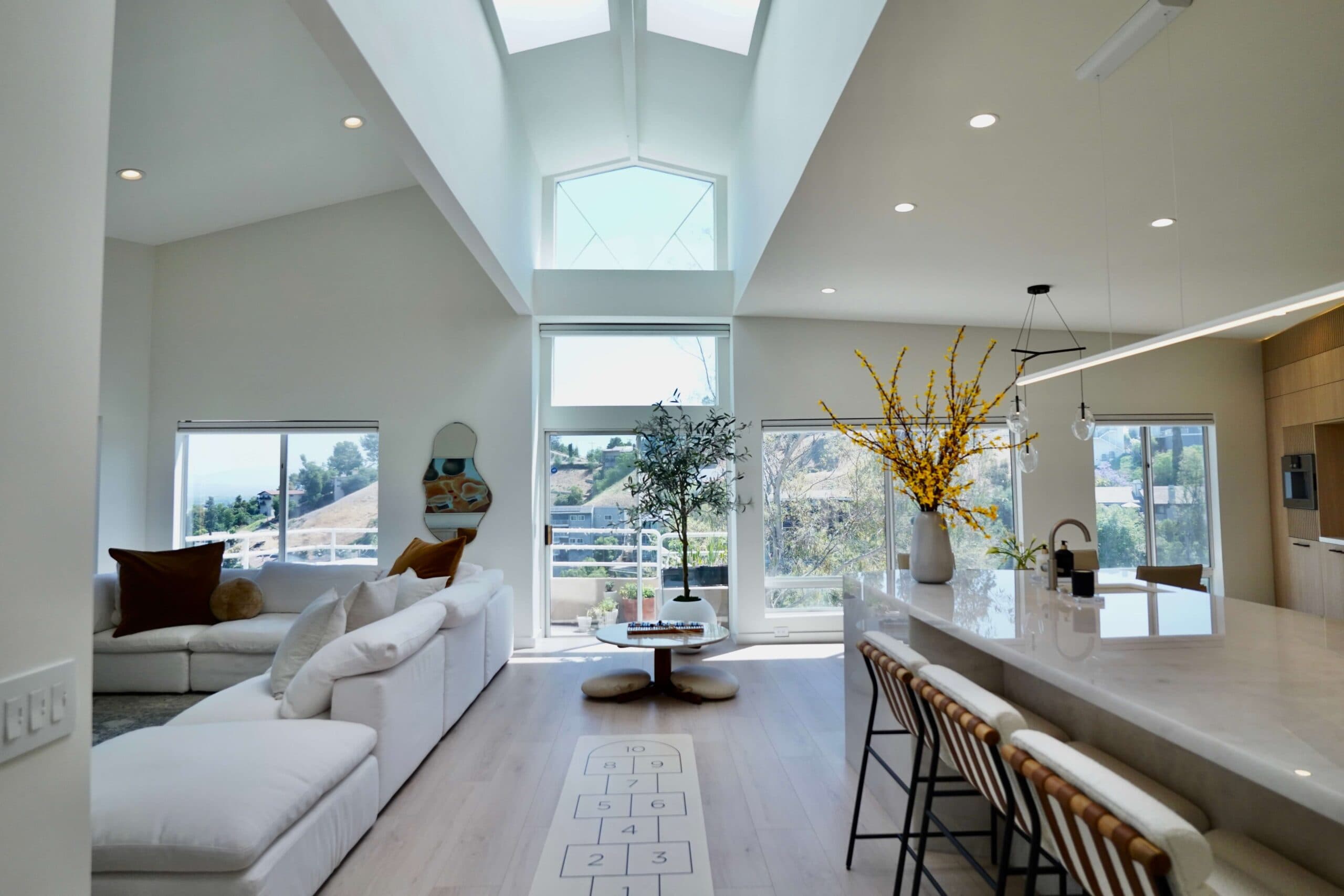 A luxury open-concept living space with vaulted ceilings and skylights, white sectional sofa with brown throw pillows, round coffee table, potted tree, and a kitchen island with bar stools and modern chandelier.