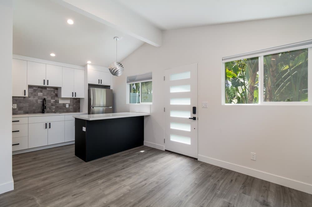 A modern minimalist ADU interior with white shaker cabinets, light gray wood-look flooring, dark gray tile backsplash, stainless steel appliances, kitchen island with black base, and a frosted glass front door.