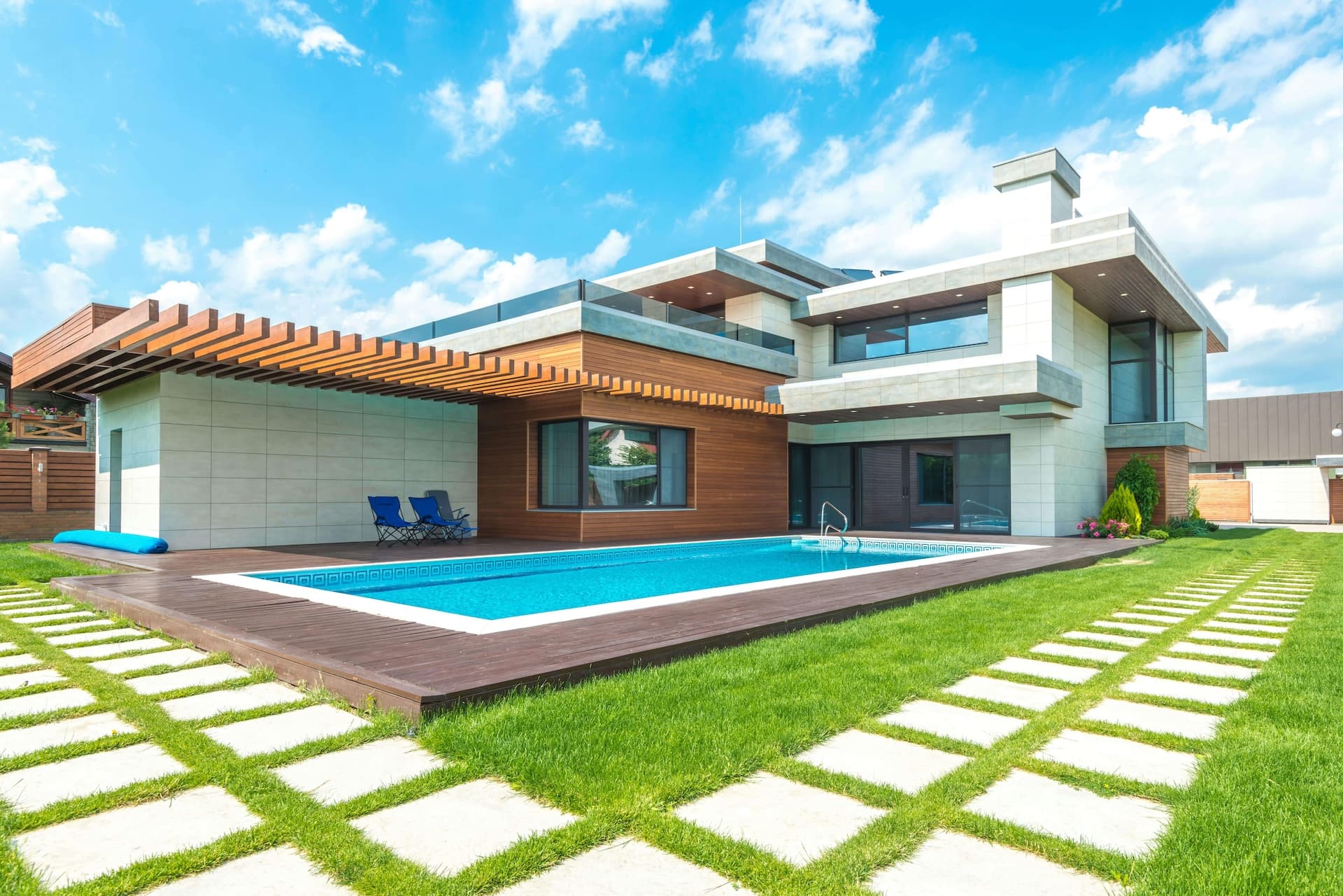 A modern outdoor living space with a rectangular swimming pool, wooden deck, blue folding chairs, lush green lawn with geometric paver patterns, and a contemporary two-story house with large glass windows.