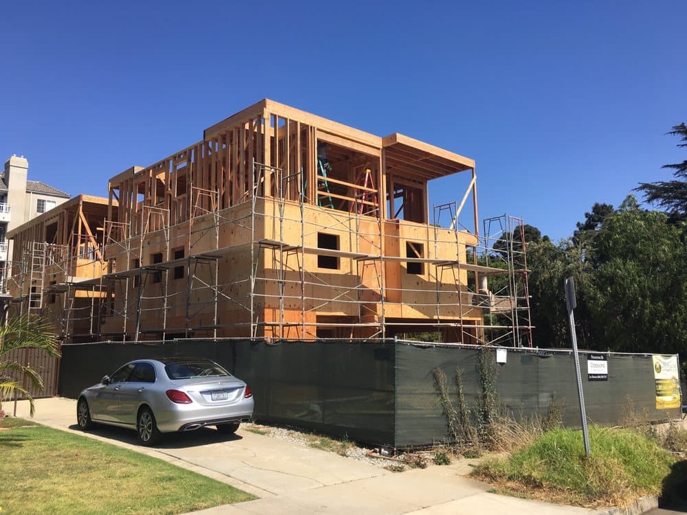 A multi-story residential building in the framing stage with wooden structural frame, extensive metal scaffolding, black safety fence, and a silver sedan parked in the driveway on a sunny day.