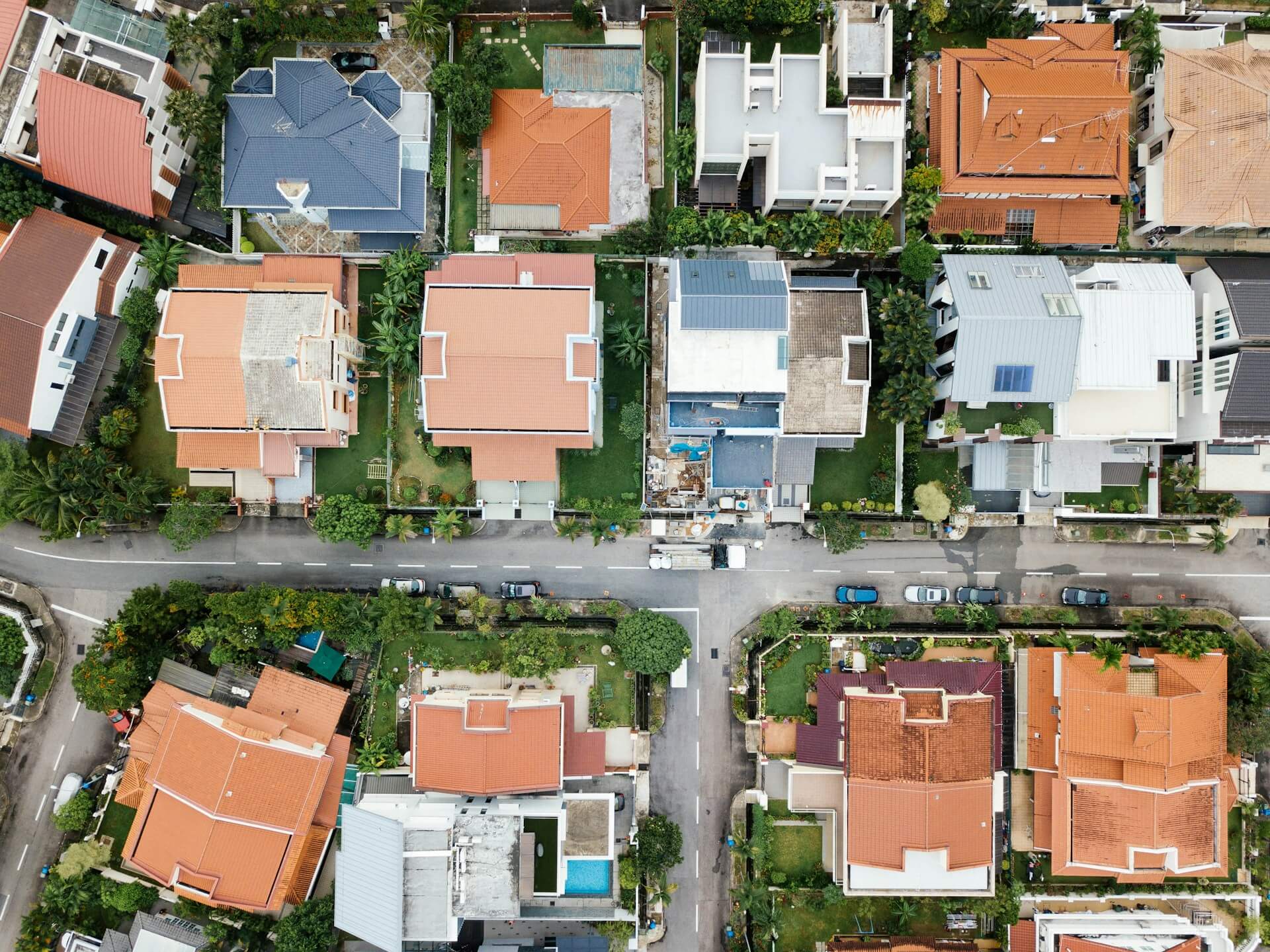 An aerial view of a well-maintained suburban residential neighborhood with houses featuring various roof colors including orange/red tiles, blue, gray and white, green lawns, trees, and streets with parked vehicles.