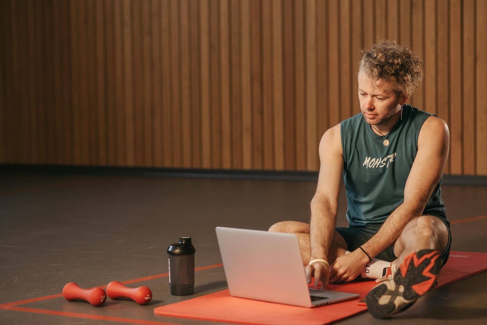 A person working on a laptop in a fitness studio with a red exercise mat, dumbbells, and wooden paneling background
