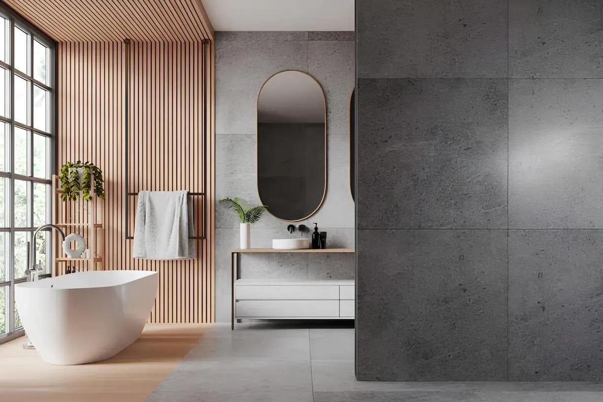 A contemporary bathroom with a white freestanding bathtub, wall-mounted vanity with vessel sink, gold-framed mirror, gray concrete-look tiles, vertical wooden slat accent wall, and floor-to-ceiling window.