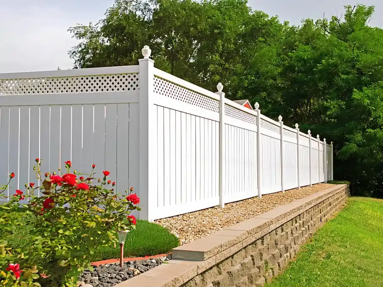 A clean white vinyl privacy fence with decorative lattice top and finial posts, mounted on a raised stone retaining wall with gravel base, set against lush green trees and a garden with red roses.