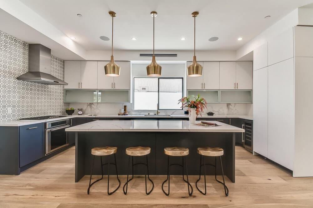 A modern, spacious kitchen with a white marble island, blue lower cabinets, gold pendant lights, stainless steel appliances, and a geometric tile backsplash. Wooden stools and hardwood flooring complete the contemporary design.