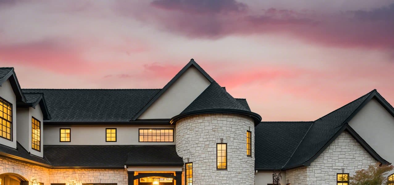 Elegant modern house with dark shingled roof and light stone facade illuminated at dusk against pink and purple sunset sky.