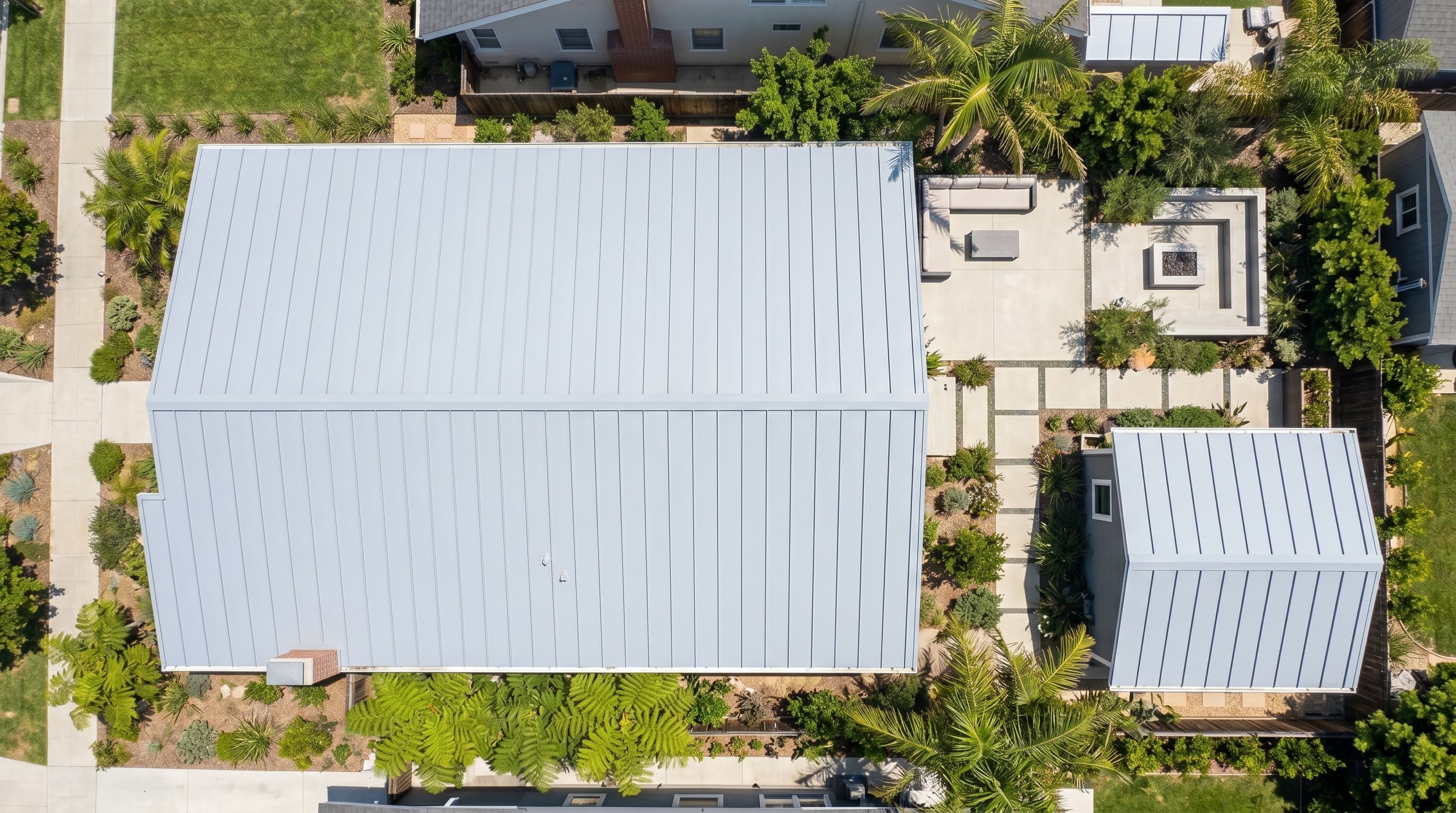 top-down drone photograph of a modern Los Angeles home with a light gray reflective standing seam metal roof and palm trees.